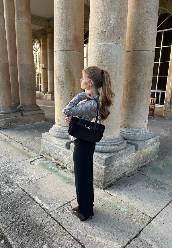 Une femme portant un sac à main noir se tient parmi de hautes colonnes en pierre, devant un majestueux bâtiment aux fenêtres cintrées.