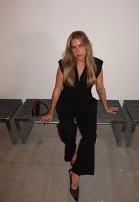 Svart ärmlös jumpsuit med djup V-ringning, kombinerad med genomskinliga svarta klackar. Sitter på reflekterande metallbänkar med en burgundy handväska.