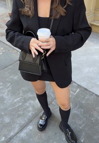 Črn oversize blazer preko črnega topa brez rokavov in mini krila, v kombinaciji z črnimi nogavicami do kolen ter sijajnimi črnimi loaferji. Drži skodelico kave.