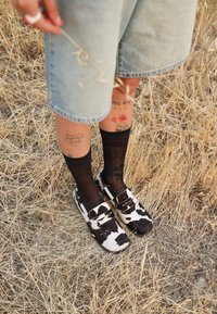 Pantofi slip-on cu imprimeu de vacă, cu accente negre, pantaloni scurți din denim și șosete negre până la genunchi, stând pe iarbă uscată și pământ.
