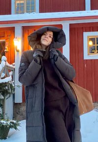 Manteau matelassé gris foncé avec une grande capuche, tissu texturé, poches latérales et intérieur chaud, assorti à un sac marron et des gants d'hiver.