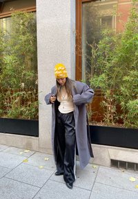 Manteau long gris, chemise beige à manches longues, pantalon de jogging noir avec des rayures blanches, chaussures noires et bonnet jaune avec logo, se tenant près de la verdure.