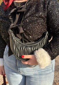 Sac à main en cuir noir matelass é avec une forme incurvée, une quincaillerie argentée et une bandoulière en chaîne, tenu contre un haut noir texturé orné de sequins.