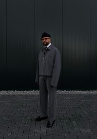 Γκρι κοντό μπουφάν με φερμουάρ και μπροστινές τσέπες, συνδυασ�μένο με ταιριαστά παντελόνια. Μαύρα δερμάτινα παπούτσια και μαύρο σκουφάκι. Ενιαίος φόντος.