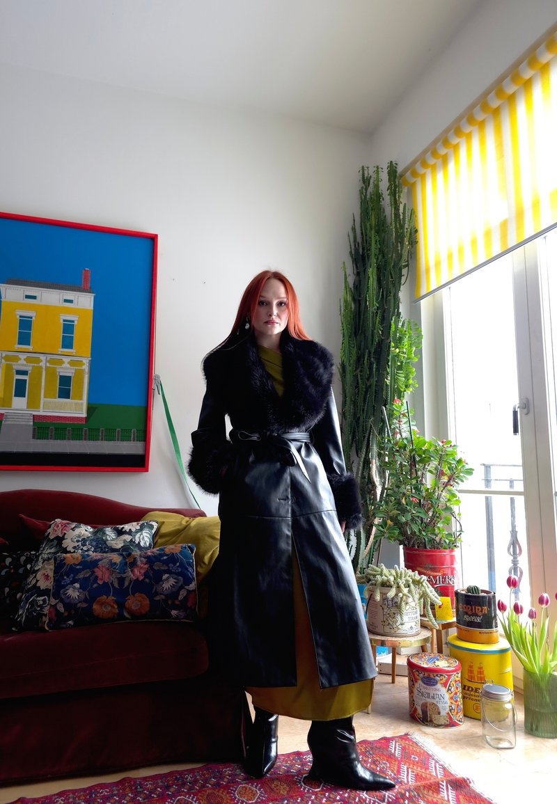 Femme aux cheveux roux portant un long manteau noir bordé de fourrure, debout dans une pièce lumineuse avec des plantes, des coussins colorés et un store à rayures jaunes.