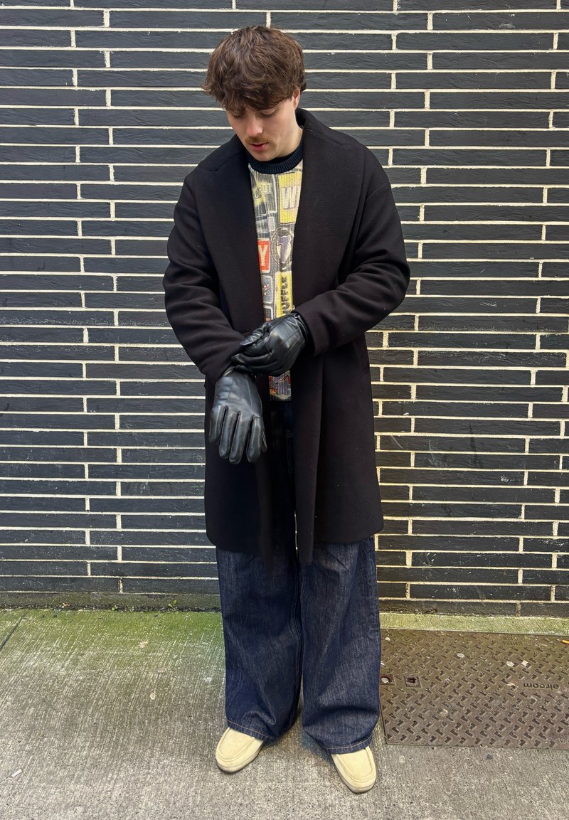 Jonge man in een zwarte jas en handschoenen die een handschoen aanpast, draagt brede donkere jeans en lichte schoenen, staand tegen een zwart-witte bakstenen muur.