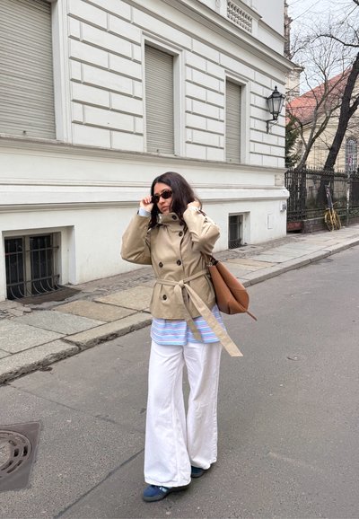 Mujer ajustándose las gafas de sol en la acera de la ciudad, vestida con un abrigo beige, pantalones anchos blancos, zapatos azules y llevando un bolso marrón junto a un edificio blanco.