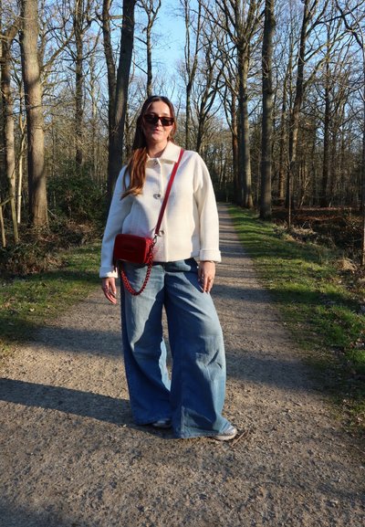 Chaqueta blanca de textura con cuello, vaqueros de pierna ancha azules y un bolso cruzado rojo con cadena, situado en un sendero natural.