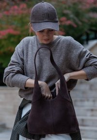 Rjava semiš torbica s ukrivljeno obliko, širokim ramenskim trakom in vidnimi šivi. Mehak videz in neopazna eleganca.