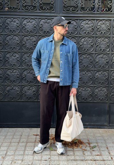 Camisa de denim con dos bolsillos en el pecho, suéter de punto verde oliva, pantalones negros, zapatillas grises y blancas, y una bolsa de tejido color crema. Fondo negro mate.