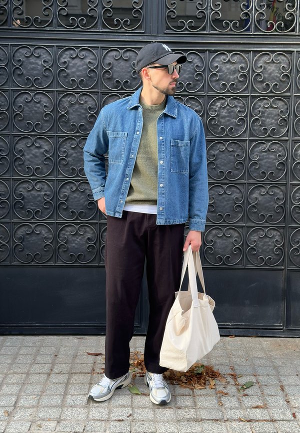 Denimskjorte med to brystlommer, olivengrøn striktrøje, sorte bukser, grå og hvide sneakers samt en crèmefarvet totebag. Mat sort baggrund.