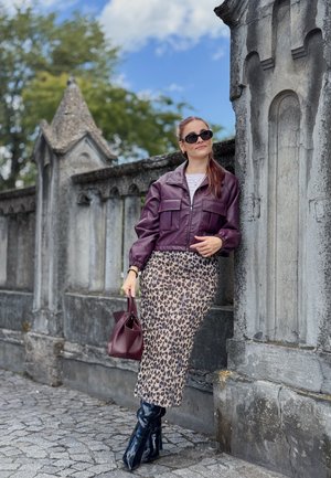 Chaqueta de cuero púrpura corta con dos bolsillos frontales, combinada con una falda midi de estampado de leopardo y botas negras hasta la rodilla. Sosteniendo un bolso burdeos.