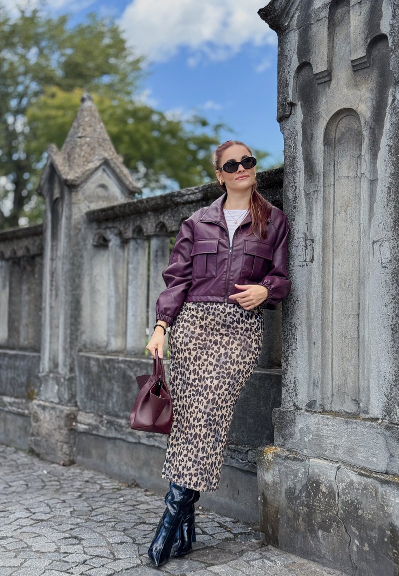 Veste cropped en cuir violet avec deux poches avant, associée à une jupe midi à imprimé léopard et des bottes noires à hauteur de genou. Tenant un sac à main bourgogne.