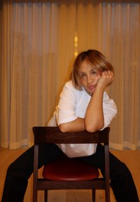 Sedia di legno con un cuscino rosso. Persona in camicia bianca posa con le braccia appoggiate allo schienale, davanti a uno sfondo di tende trasparenti.
