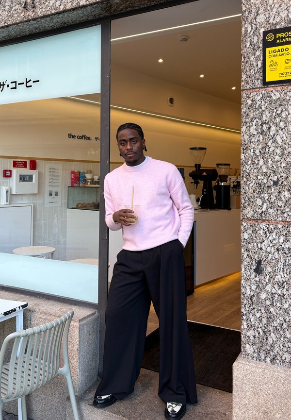 Hombre con suéter rosa y pantalones anchos negros está en la puerta del café sosteniendo una bebida con pajita, junto a una silla y mesa exteriores.