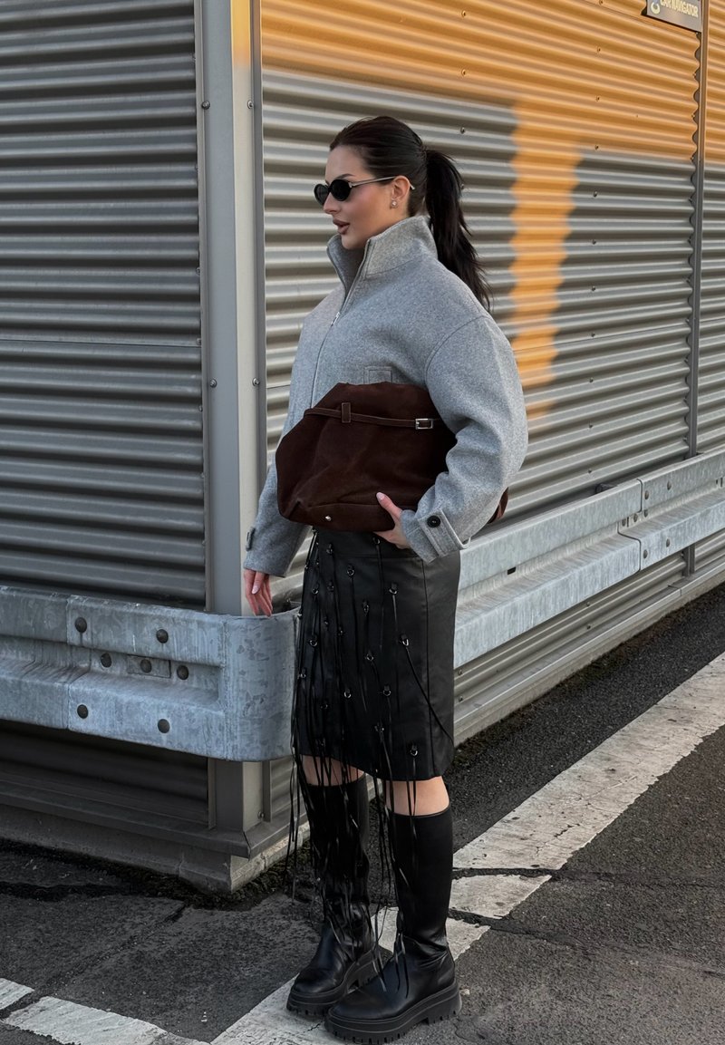 Femme avec queue de cheval brune portant une veste grise zippée, une jupe en cuir noire à franges, des bottes noires et des lunettes de soleil, tenant un grand sac marron en plein air.