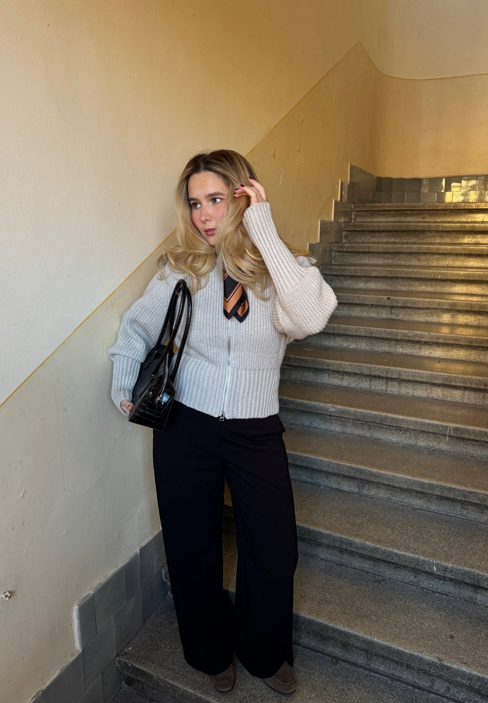 Cardigan gris clair en maille avec texture côtelée, pantalon noir à jambes larges et cravate marron rayée, accompagné d'un sac à main noir, sur des escaliers.