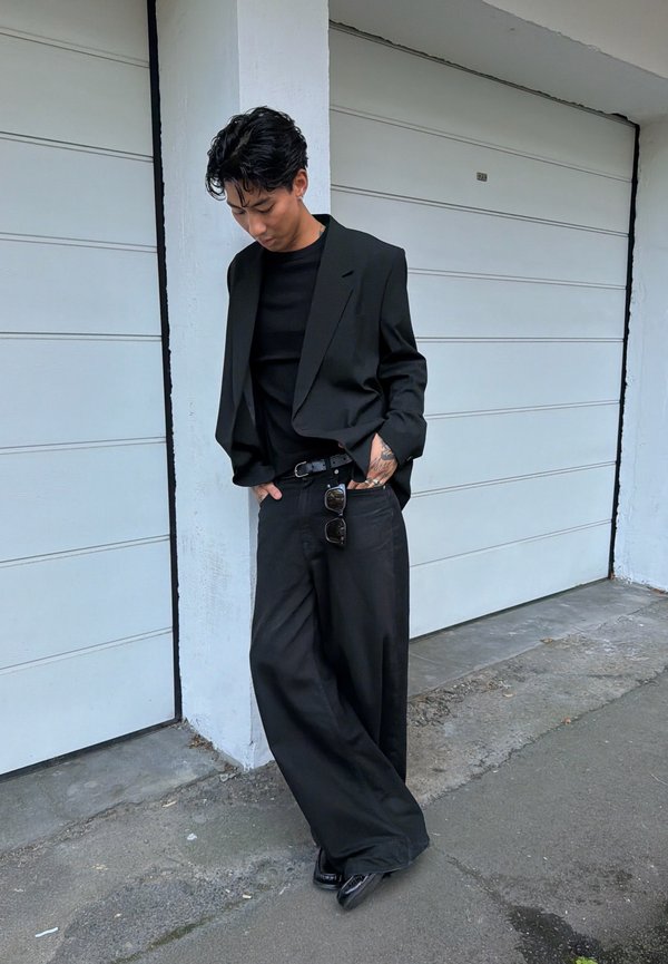 Hombre con cabello negro que lleva pantalones negros anchos, blazer negro y zapatos negros, de pie y mirando hacia abajo junto a una puerta de garaje blanca al aire libre.