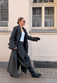 Γυναίκα που περπατάει σε πεζοδρόμιο πόλης, φορώντας μακρύ σκούρο παλτό, φούστα με ρίγες, λευκό crop top, μαύρες μπότες, γυαλιά ηλίου και κρατώντας μαύρη τσάντα.