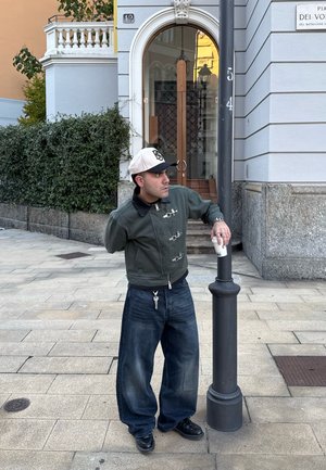 Green bomber jacket with black collar, oversized dark blue jeans, black shoes, and white baseball cap. Holds a cup near a lamp post.