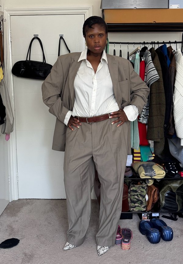 Personne se tient avec assurance dans un costume beige oversize avec une chemise blanche, des chaussures pointues en peau de serpent, dans une pièce où des vêtements et des sacs sont visibles en arrière-plan.