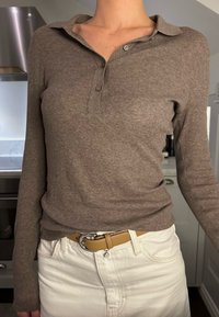 Καφέ μακρυμάνικη πουκαμίσα με γιακά, δύο κουμπιά και ριγωτή υφή, συνδυασμένη με λευκά τζιν και καφέ ζώνη με ασημένιο κούμπωμα.