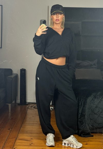 Mujer vistiendo un cropped negro, pantalones de chándal negros y zapatillas blancas, haciéndose un selfie en el espejo en una habitación con suelo de madera y muebles negros.