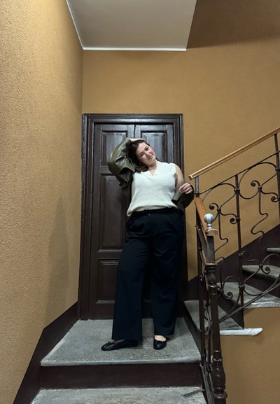 Una persona con una blusa de punto color crema y pantalones de pierna ancha negros posa junto a una puerta de madera en un vestíbulo con una pared marrón y una barandilla de hierro.