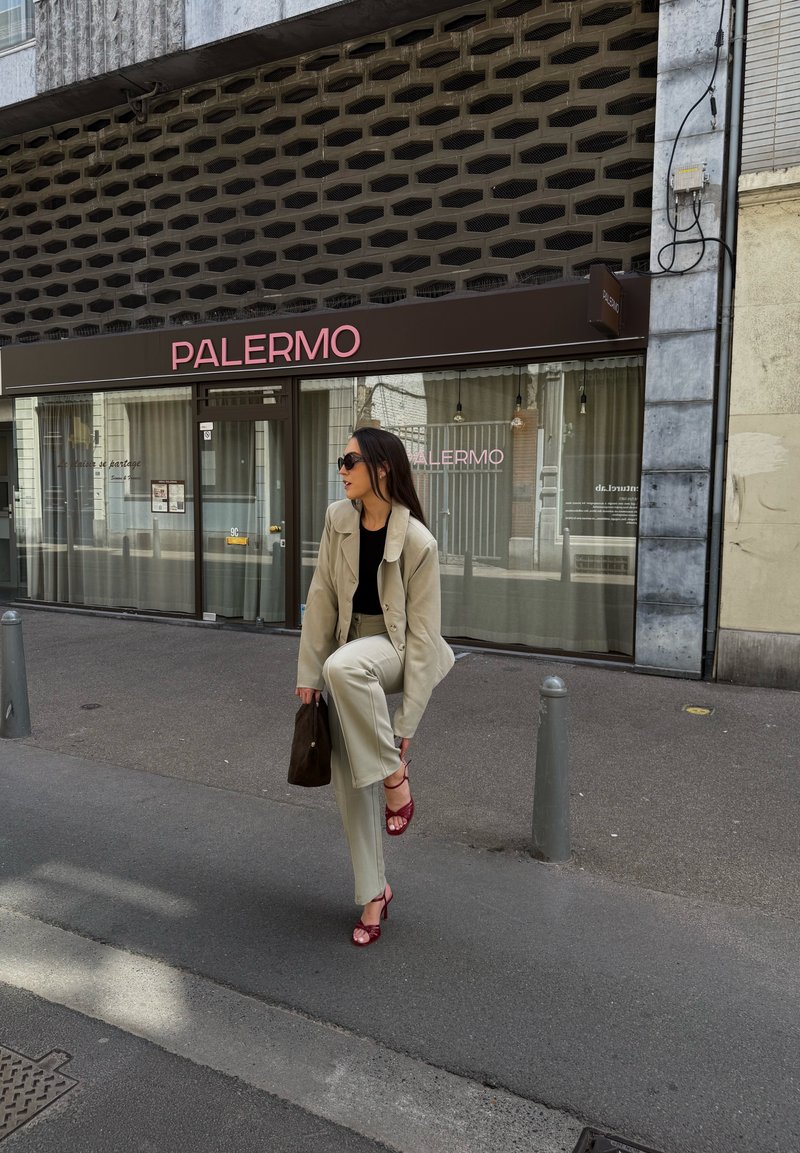 Femme en costume beige et talons rouges ajuste sa chaussure tout en se tenant sur le trottoir urbain devant un café nommé Palermo.