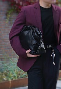 Μαύρη δερμάτινη τσάντα με ρυτιδωτό σχέδιο, κρατώντας τη κάτω από ένα βυσσινί σακάκι. Ένα απλό μαύρο πουκάμισο και κομψό μαύρο παντελόνι ολοκληρώνουν την εμφάνιση.