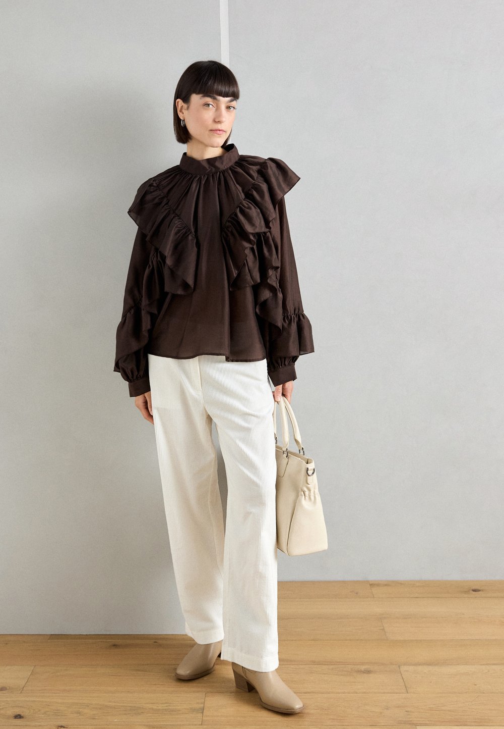 Donkerbruine blouse met rimpelaccenten, lange mouwen, hoge hals; gecombineerd met witte wijde broek en een lichtbeige handtas.