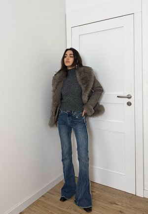 Mujer con chaqueta gris forrada de piel, suéter gris de cuello alto, vaqueros acampanados azules y zapatos negros, de pie frente a una puerta blanca y una pared blanca.