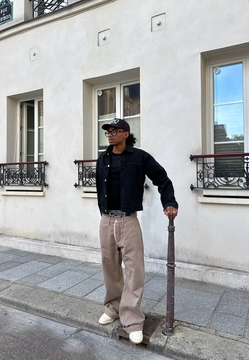 Uomo con giacca nera, pantaloni beige e cappellino in piedi sul marciapiede, appoggiato a un palo di metallo, davanti a un edificio chiaro con finestre.