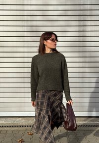 Ciemnozielony dzianinowy sweter z okrągłym dekoltem, zestawiony z plisowaną spódnicą w kratę z falbaną na dole. Dopełnieniem stylizacji jest brązowa torebka.