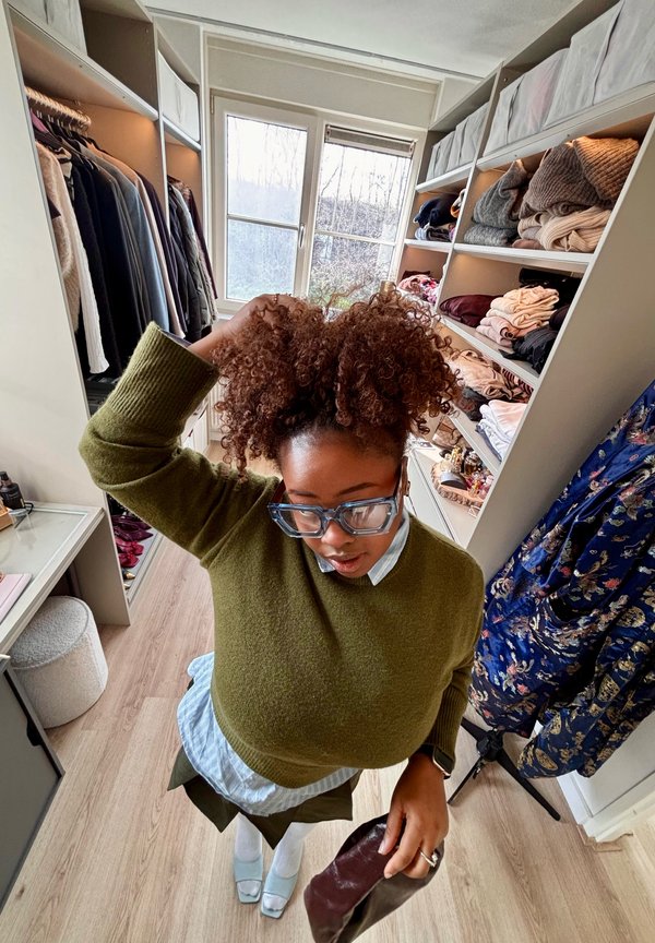 Donna che sistema i capelli ricci in una cabina armadio con scaffali di vestiti piegati, abiti appesi e una finestra che mostra una vista esterna.