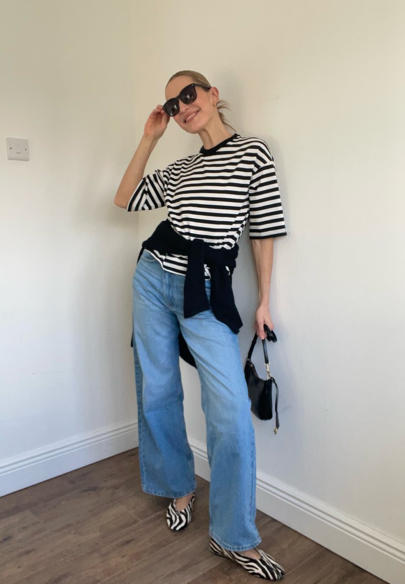 Femme portant un t-shirt rayé noir et blanc, un jean bleu clair, des chaussures à imprimé zèbre, des lunettes de soleil noires, tenant un sac à main noir, debout à l'intérieur.