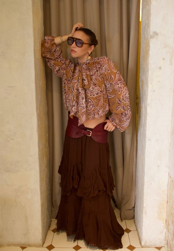 Femme portant des lunettes de soleil surdimensionnées, un chemisier marron à motifs, une large ceinture bordeaux et une longue jupe marron foncé à volants, debout entre deux murs.