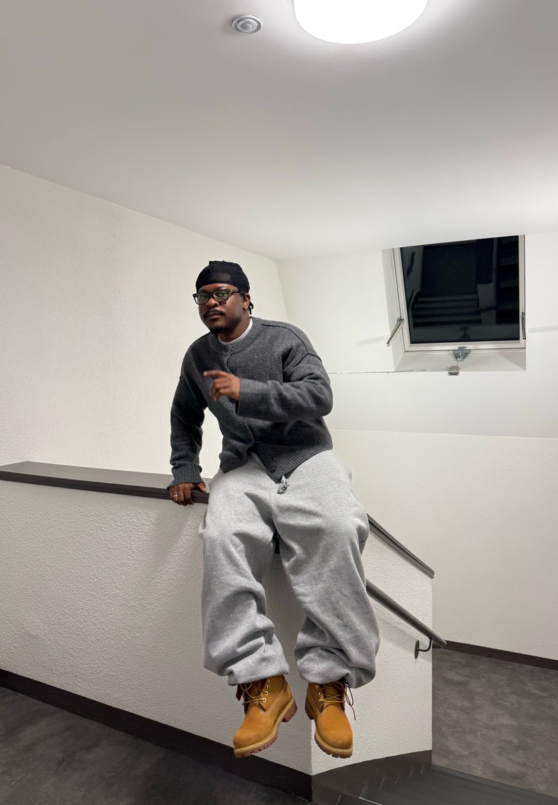 Homme portant un pull gris, un pantalon de survêtement gris ample et des bottes couleur sable, assis sur le rebord d'un escalier à l'intérieur près d'une fenêtre la nuit.