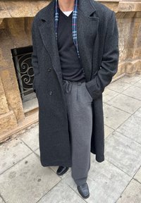 Homme portant un pardessus gris foncé, un pull noir, un pantalon gris et des chaussures noires, debout dans une rue pavée en pierre près d'un mur en pierre.