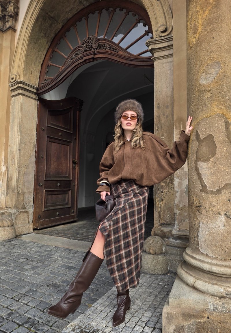 Maglia oversize in camoscio marrone, gonna midi a quadri con spacco e stivali marroni al ginocchio. Accessori includono cappello di pelliccia e occhiali da sole sfumati.