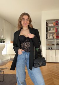 Jeune femme portant un haut en dentelle noire, un blazer noir oversized, un jean bleu clair, tenant un sac à main Prada noir dans un salon moderne.