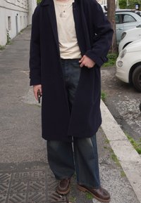 Casaco de marinha sobre uma blusa bege, jeans de perna larga azul e mocassins castanhos. Detalhes notáveis incluem uma textura cinza e costura visível nos sapatos.