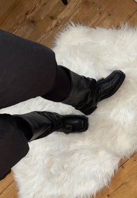 Schwarze Leder-Stiefel bis zum Knie mit einem Schnallen-Detail, getragen über schwarzen Leggings, auf einem strukturierten weißen Kunstfellteppich. Sichtbarer Holzboden.