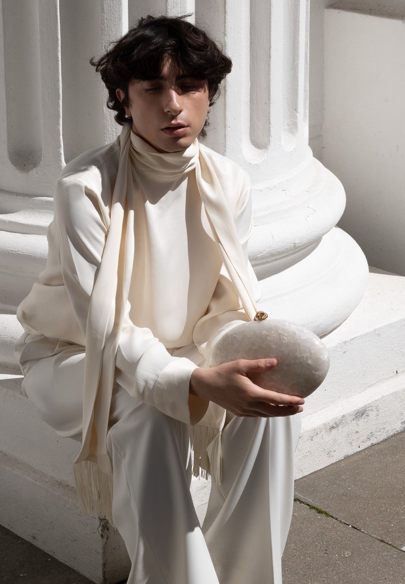 Jeune personne aux cheveux foncés porte une tenue crème et une écharpe, assise sur une base architecturale blanche, tenant une pochette ronde texturée.