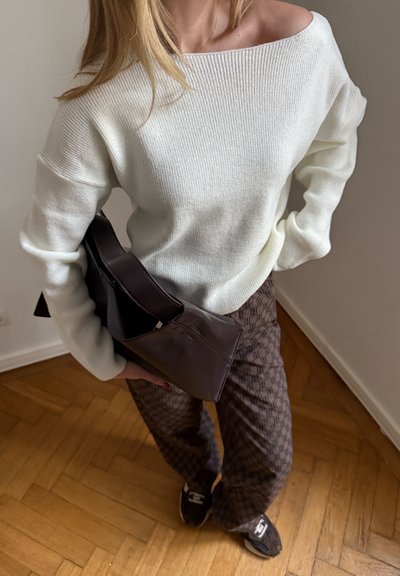 Mujer con suéter blanco de hombros descubiertos, pantalones marrones con estampado, zapatillas negras y sosteniendo un gran bolso marrón oscuro a su lado.