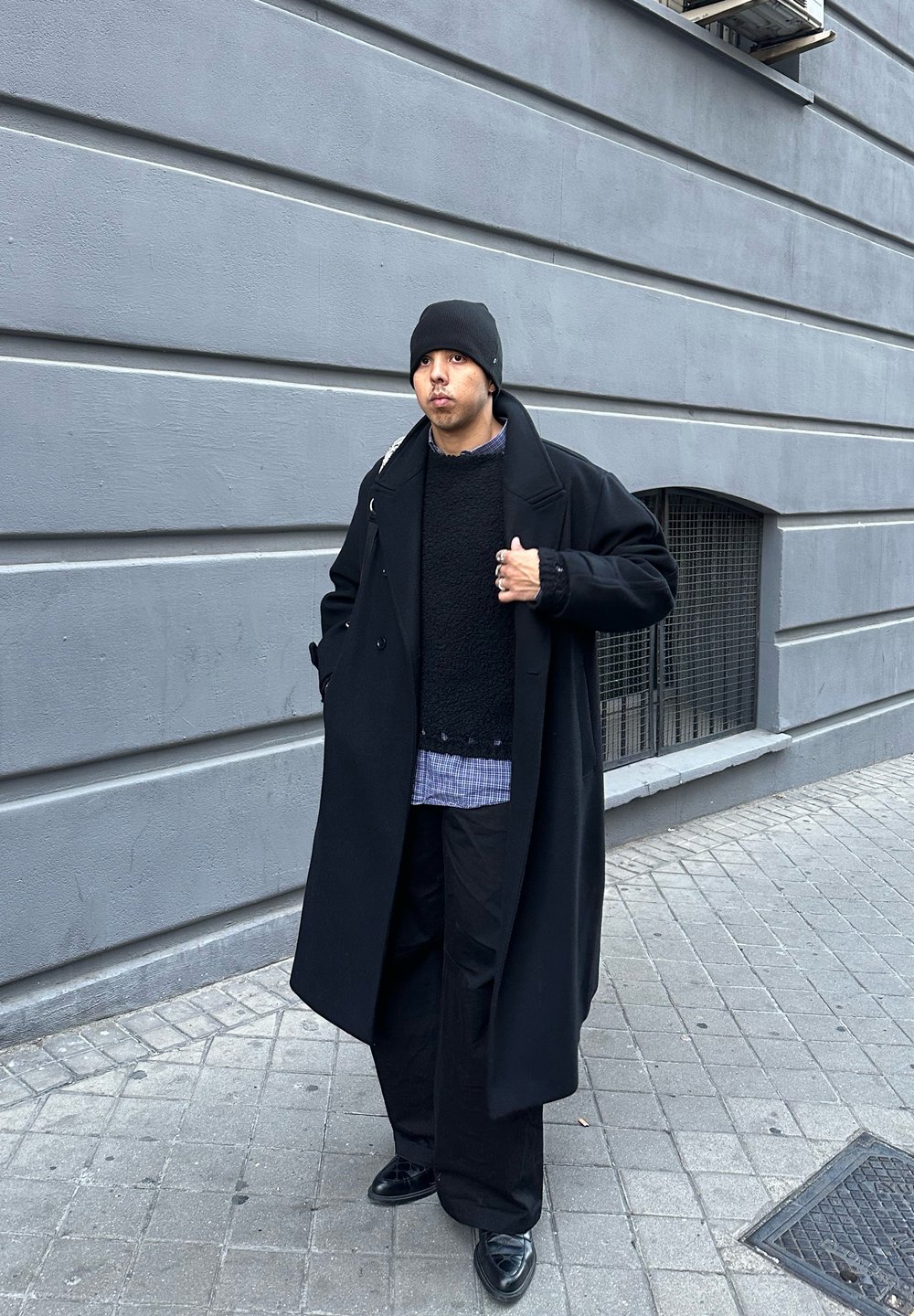 Černý kabát, tmavý strukturovaný svetr, modrá kostkovaná košile, široké černé kalhoty, černá čepice, černé boty, stojící proti šedé zdi.