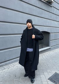 Černý kabát, tmavý strukturovaný svetr, modrá kostkovaná košile, široké černé kalhoty, černá čepice, černé boty, stojící proti šedé zdi.