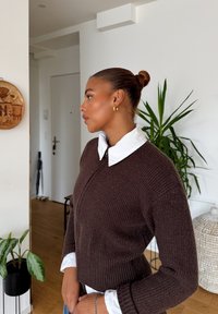 Vrouw met een bun-kapsel die gouden ringoorbellen draagt, een witte blouse onder een bruine gebreide trui, staande binnen bij planten en houten decor.