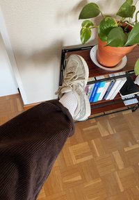 Ανοιχτό γκρι αθλητικό παπούτσι με υφασμένο πλέγμα, φορεμένο με λευκές κάλτσες. Ορατό το παντελόνι καμπούρας σε σκούρο καφέ χρώμα. Ξύλινο πάτωμα στο φόντο.