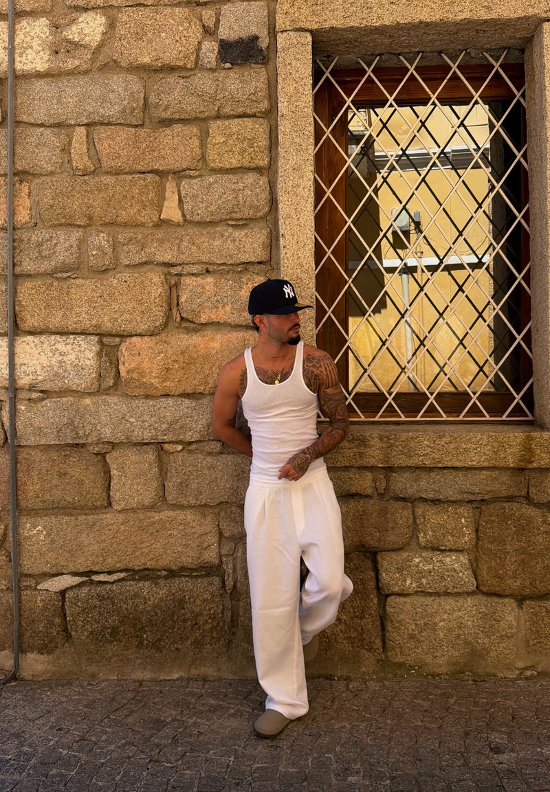 Homme tatoué portant un débardeur blanc, un pantalon et une casquette sombre, appuyé contre un mur en pierre près d'une fenêtre grillagée dans une rue pavée.
